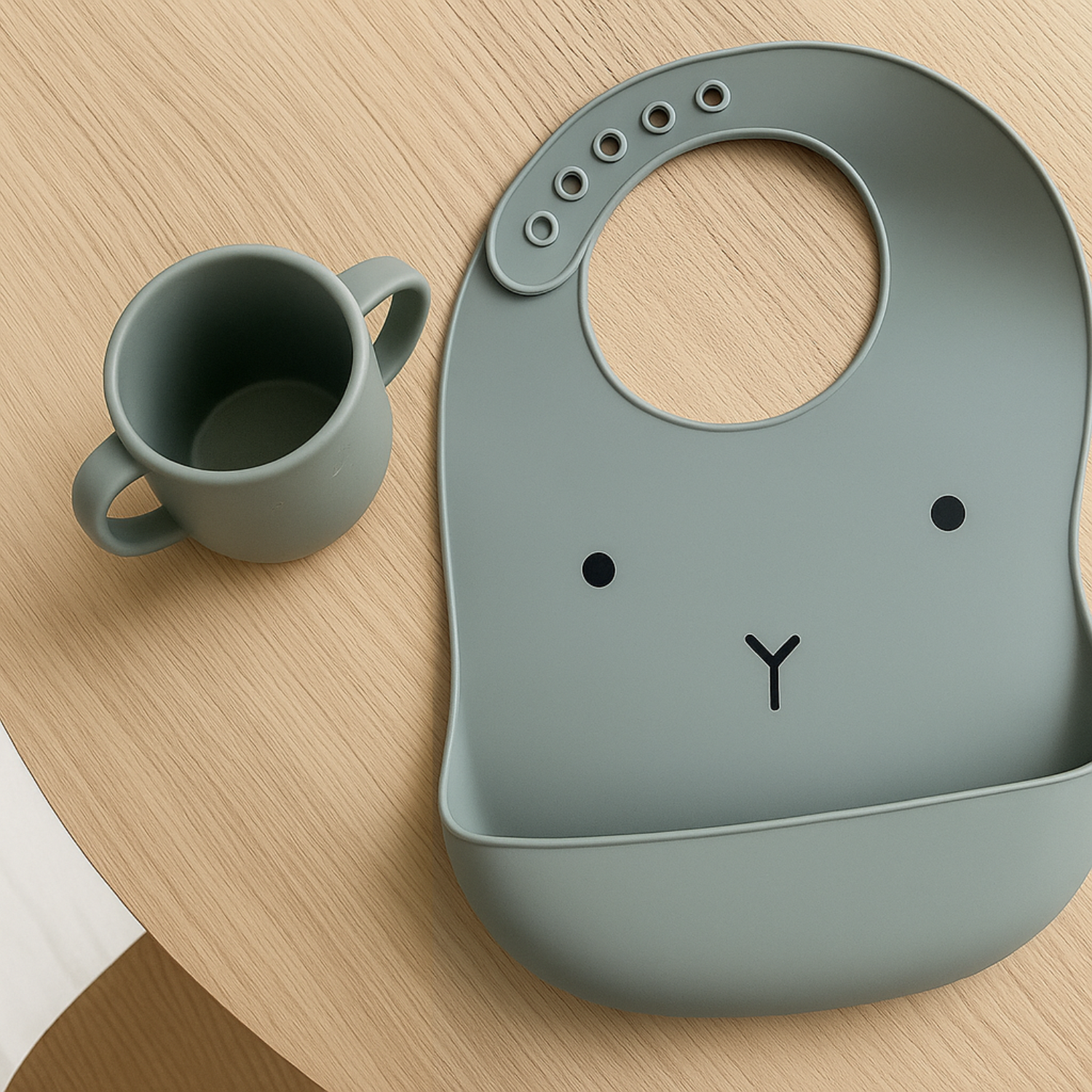 Présentation d’un bavoir en silicone pour enfants de couleur grise, avec un motif posé sur une table en bois avec une tasse à boire juste à côté.