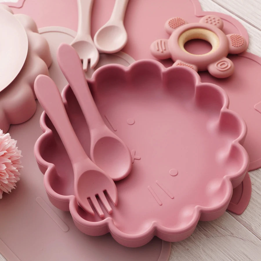 Présentation d'accessoires pour bébé en silicone rose, comprenant une assiette, des couverts et d'autres accessoires.
