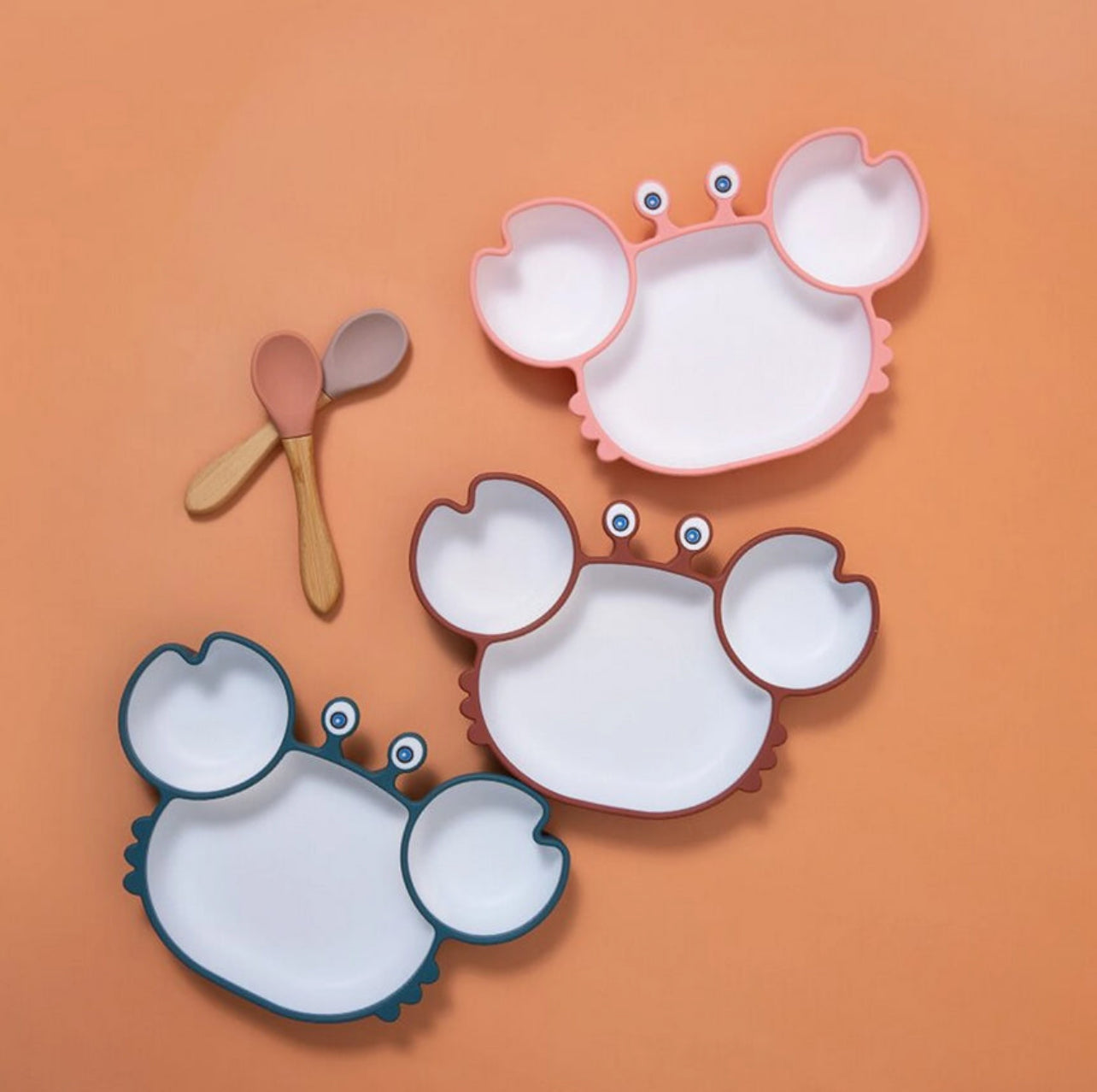 Présentation de trois assiette en forme de crabe de couleurs différente avec deux cuillère côte à côte sur un fond orange.