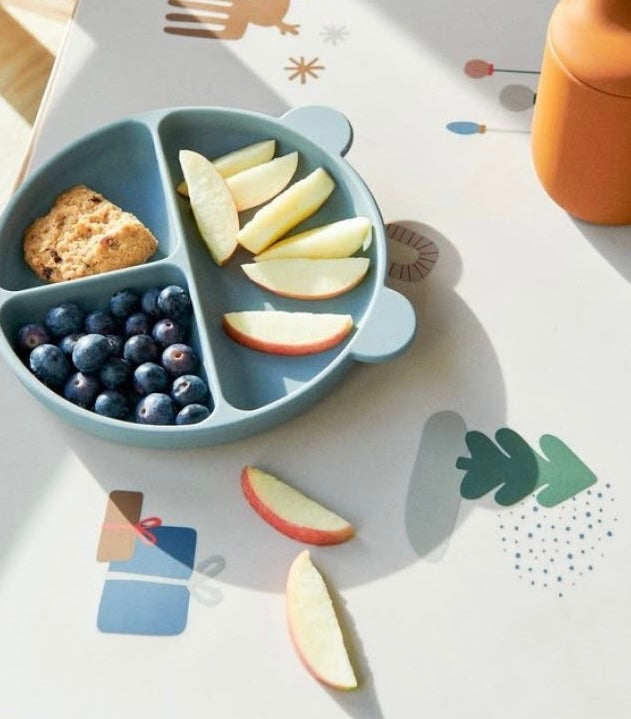 Présentation d’une assiette en forme d’ours de couleur bleu avec des aliment dedans il y a de la pomme un bout de pain et des myrtilles.
