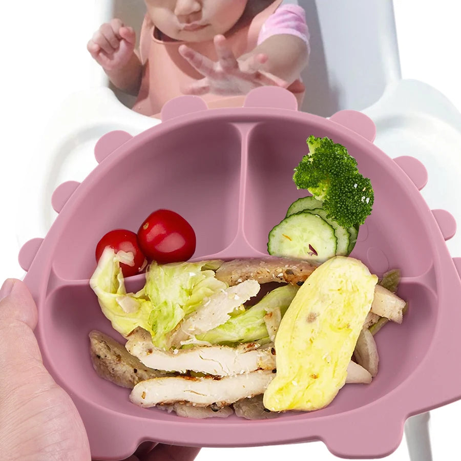 Présentation d'une assiette pour bébé rose en forme de dinosaure avec des compartiments, remplie de nourriture, avec un bébé en arrière-plan.