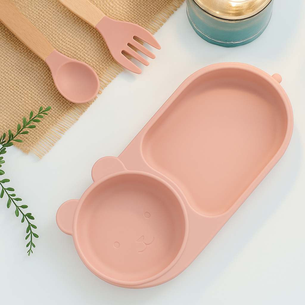 Présentation produit d’une assiette en silicone pour enfants, qui a deux compartiments une partie plus grand et une une partie qui fait bol et la partie qui fait bol a des oreilles et un visage d’ours, des couleurs roses sur un fond de table bleue avec des couverts en arrière, plan.