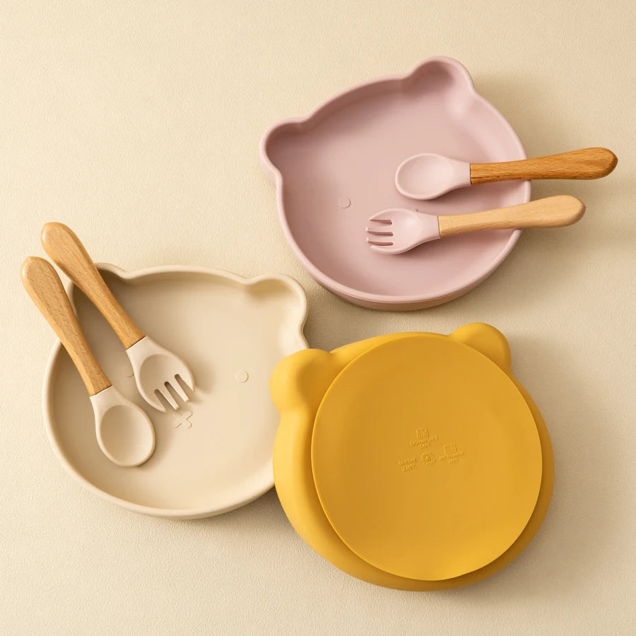 Présentation d’un kit alimentation pour bébé avec des assiettes en forme d’ours et des couverts en bois et embout silicone de différentes couleurs, jaune, beige et rose clair.