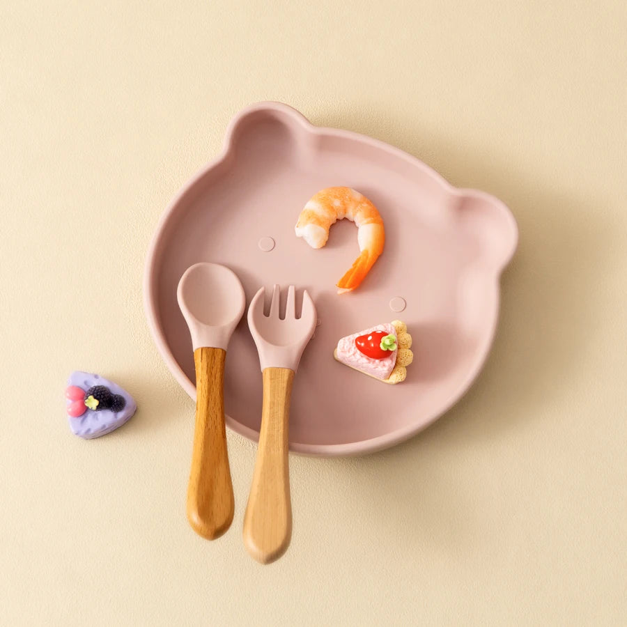 Présentation d’une assiette pour bébé en forme d’ours, rose avec des couverts en bois et silicone et des aliments disposés dessus.
