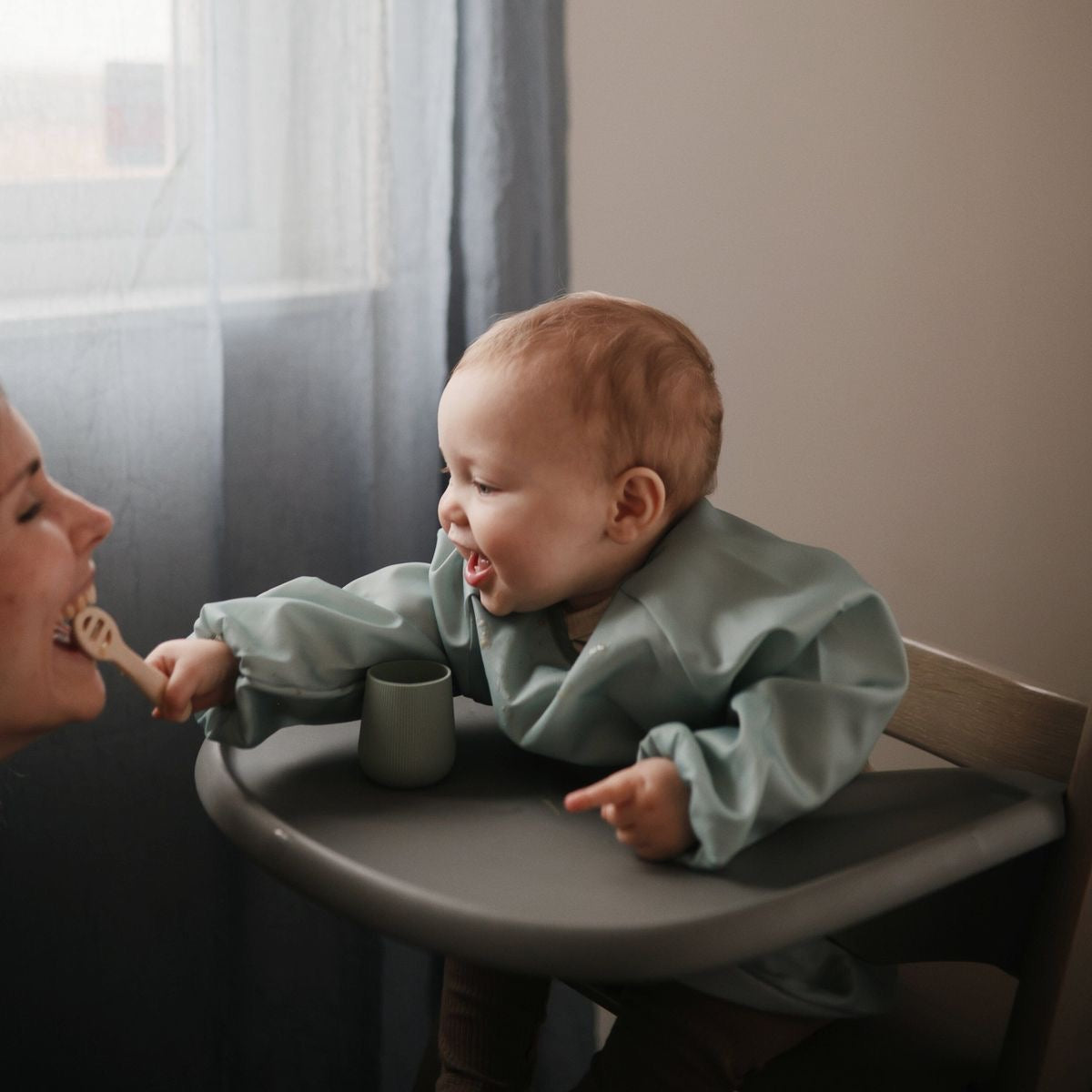 Présentation d’un bébé qui porte un bavoir à manches longues de couleur verte, qui est assis dans une chaise haute, et sourit à sa maman en lui présentant une cuillère.