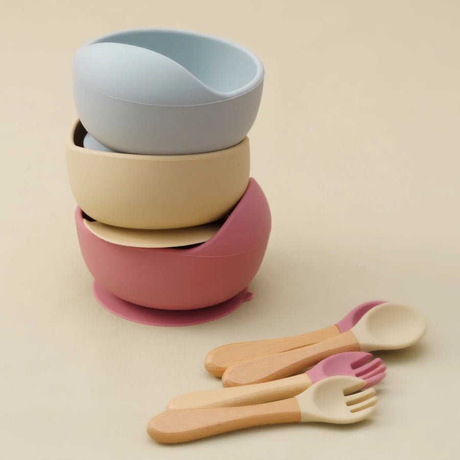 Présentation d'un ensemble de bols pour bébé en silicone empilés de différentes couleurs, avec une cuillère et une fourchette en bois.
