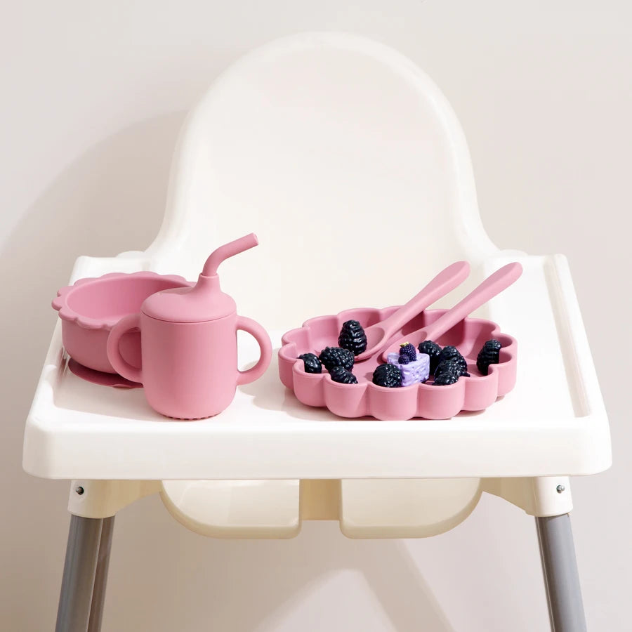 Présentation d'une chaise haute pour bébé avec un ensemble de vaisselle en silicone rose, incluant une assiette, un bol, un gobelet et des couverts.