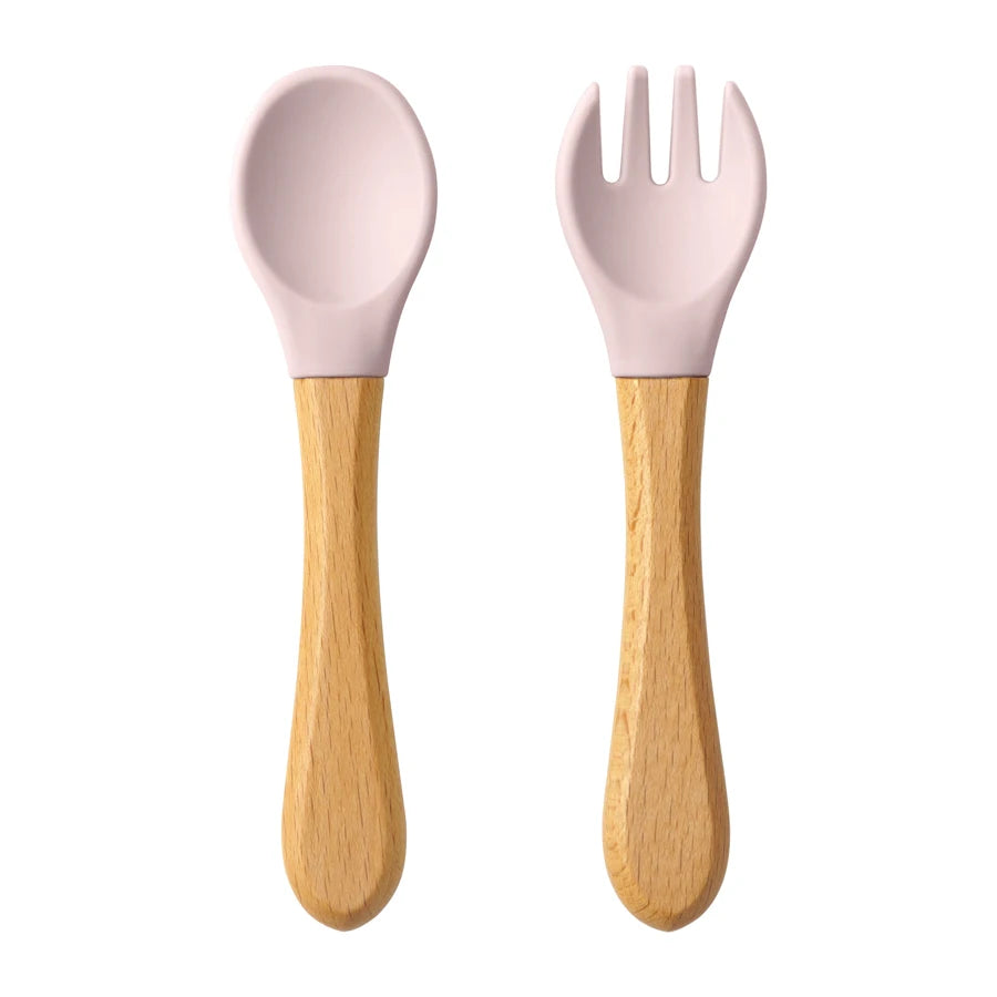 Présentation d’une fourchette et d’une cuillère pour bébé en bois avec des embouts silicone, rose clair