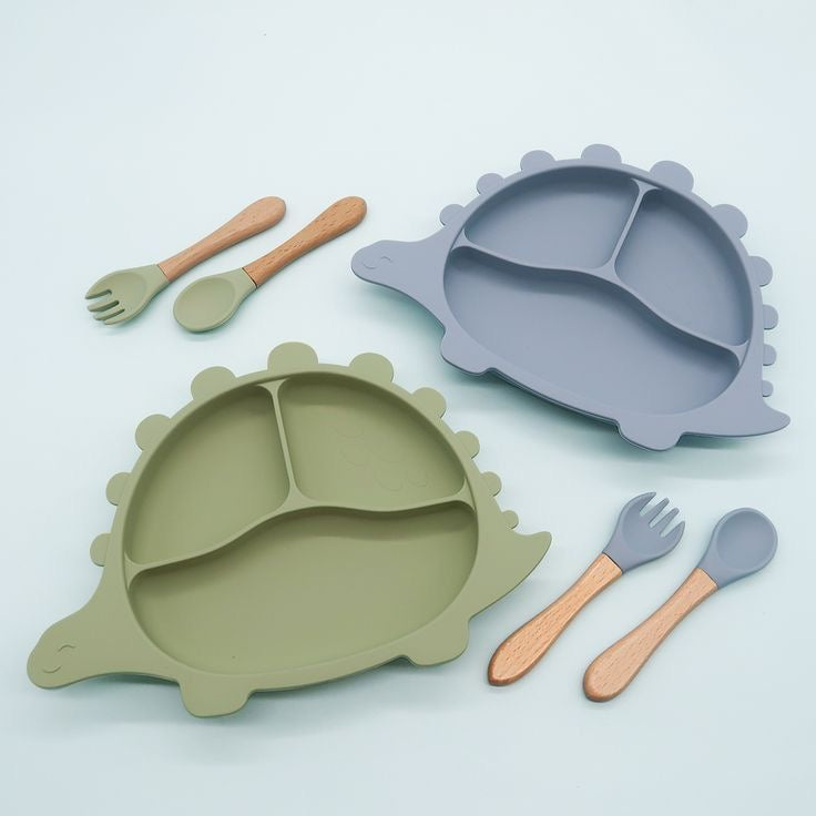 Présentation d'un ensemble de deux assiettes pour bébé en silicone en forme de dinosaure, de couleur verte et bleu, avec des couverts assortis.