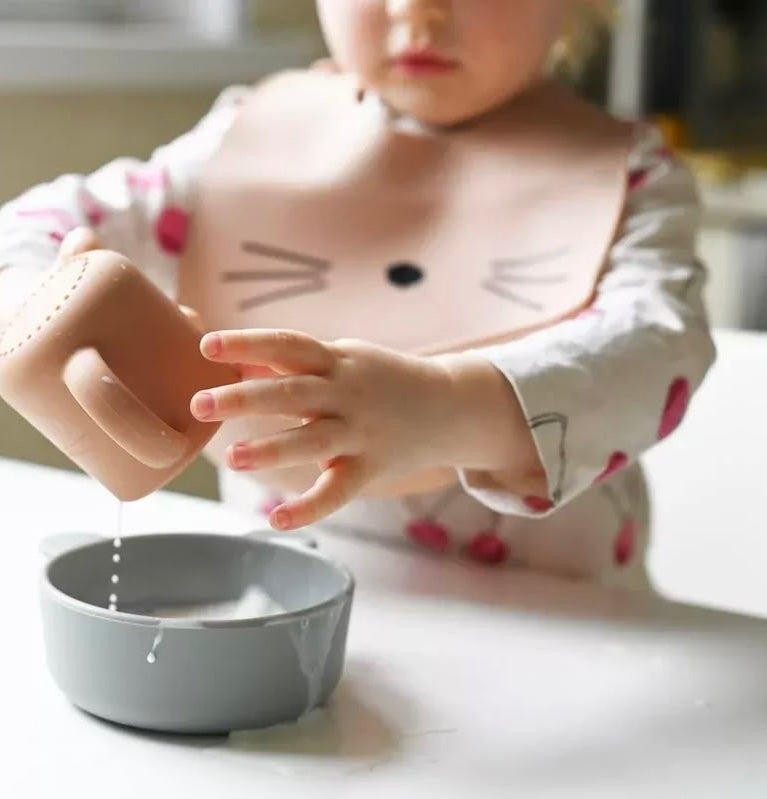 Présentation d’un enfant qui joue avec sa tasse, mais en arrière, elle porte un bavoir fantaisie avec un motif de moustache de chat, de couleur orange