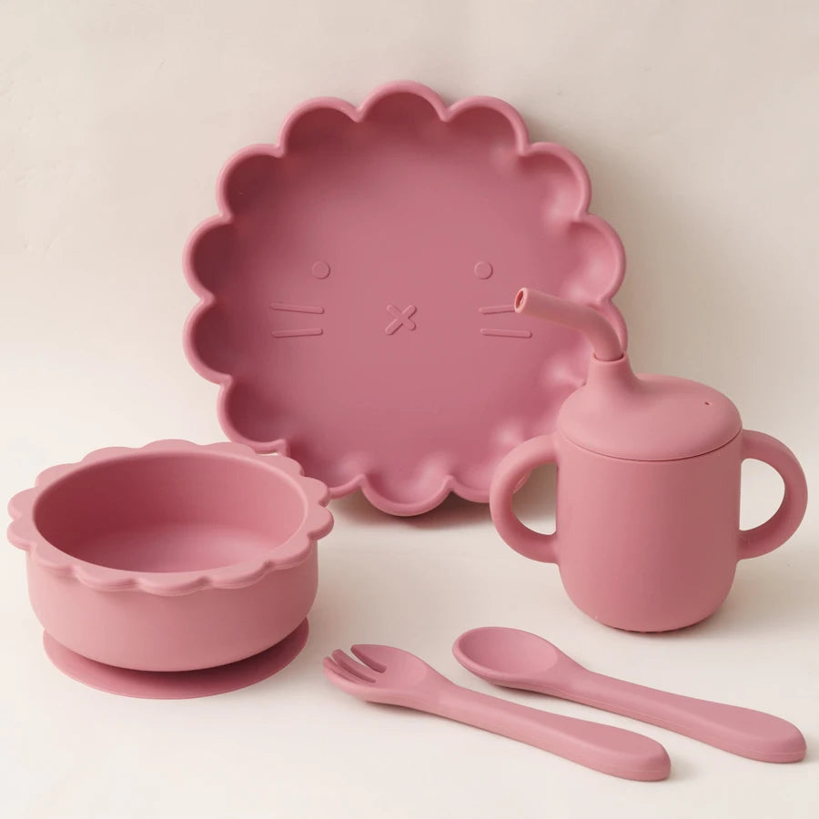 Présentation d'une assiette, d'un bol, d'un gobelet et de couverts pour bébé en silicone rose foncé, sur un fond blanc.