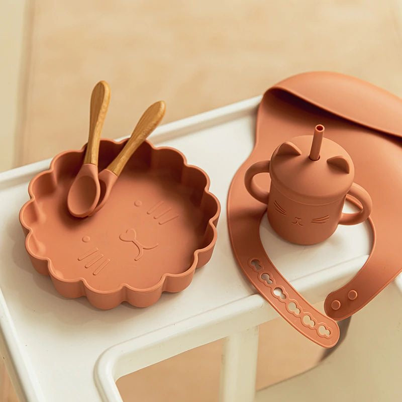Présentation d'un set repas pour bébé en silicone comprenant une assiette, une cuillère, une tasse et un bavoir, de couleur terracotta.