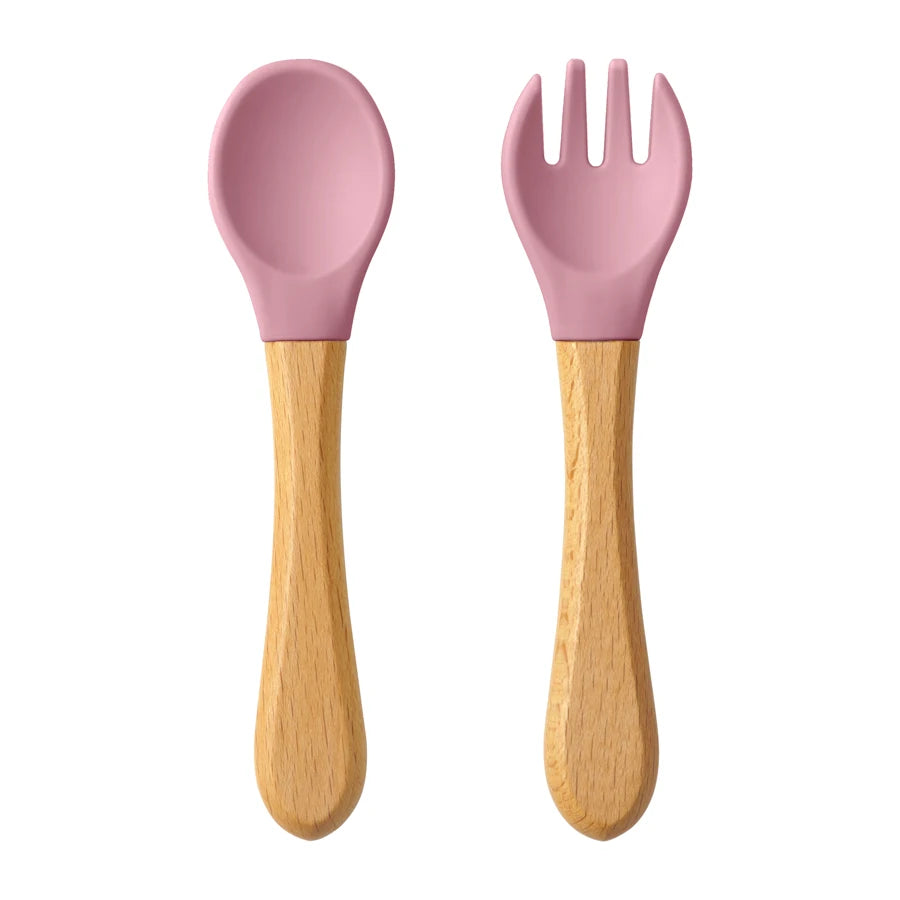 Présentation d’une fourchette et d’une cuillère pour bébé en bois avec des embouts silicone, rose.