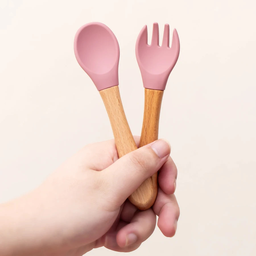 Présentation d’une fourchette et d’une cuillère pour bébé, en bois, avec des sangles en silicone, rose, tenue par une main.