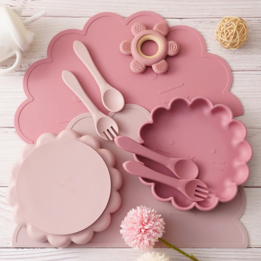 Présentation d'accessoires pour le repas de bébé en silicone rose, comprenant une assiette, des couverts, un bavoir et un anneau de dentition.